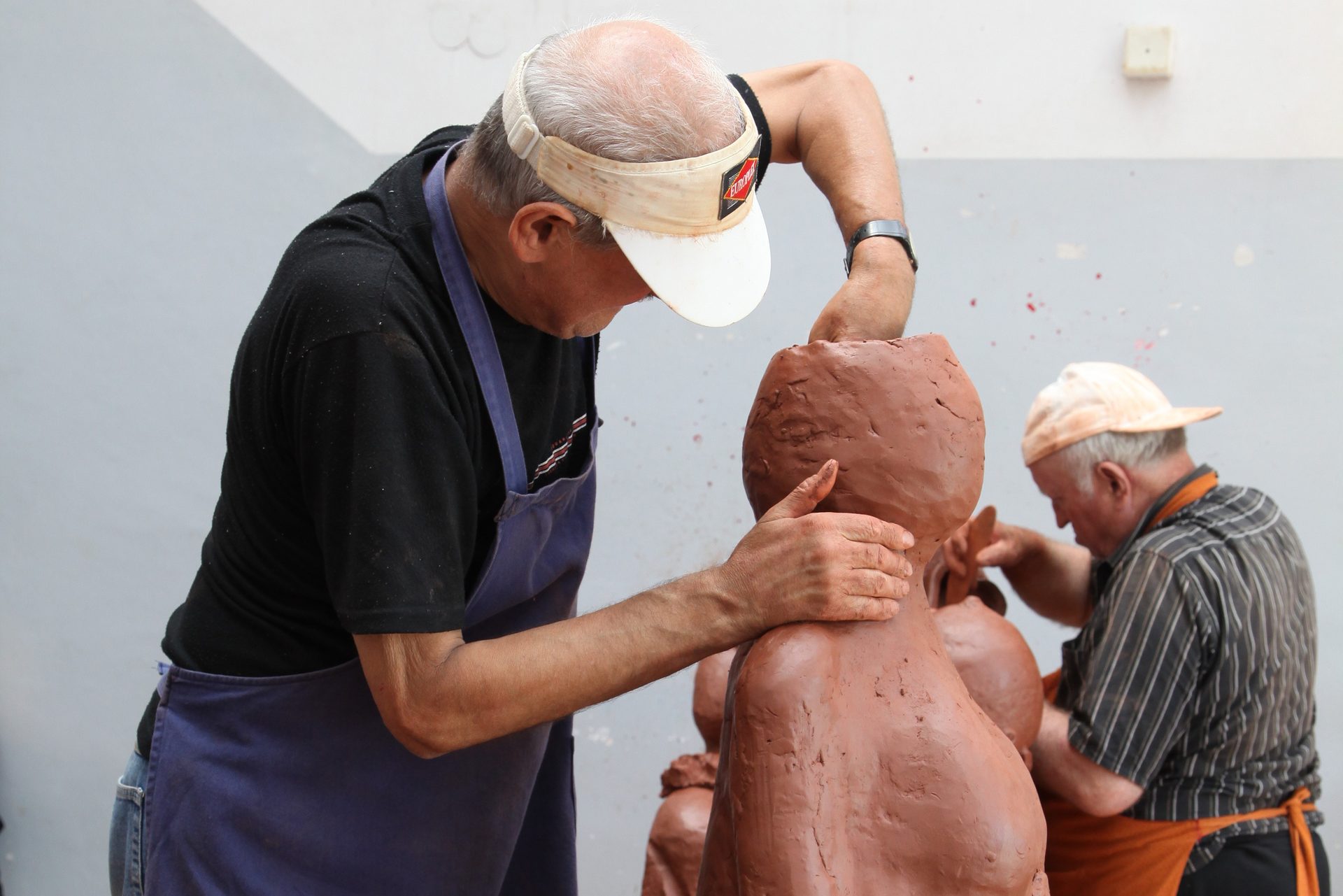 Zdjęcie przedstawia osoby znajdujące się w pracowni rzeźbiarskiej. Na pierwszym planie mężczyzna modeluje w glinie postać kobiety.