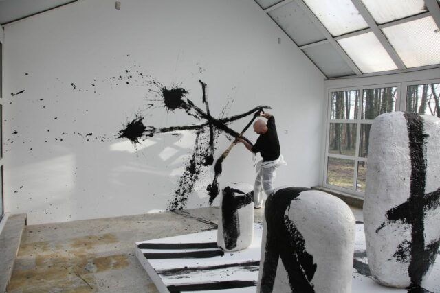 Artysta Michał Puszczyński rozchlapuje na białej ścianie czarną farbę za pomocą dużego pędzla. Na pierwszym planie ustawione pionowo ceramiczne rzeźby w formie dużych owalnych białych kamieni.